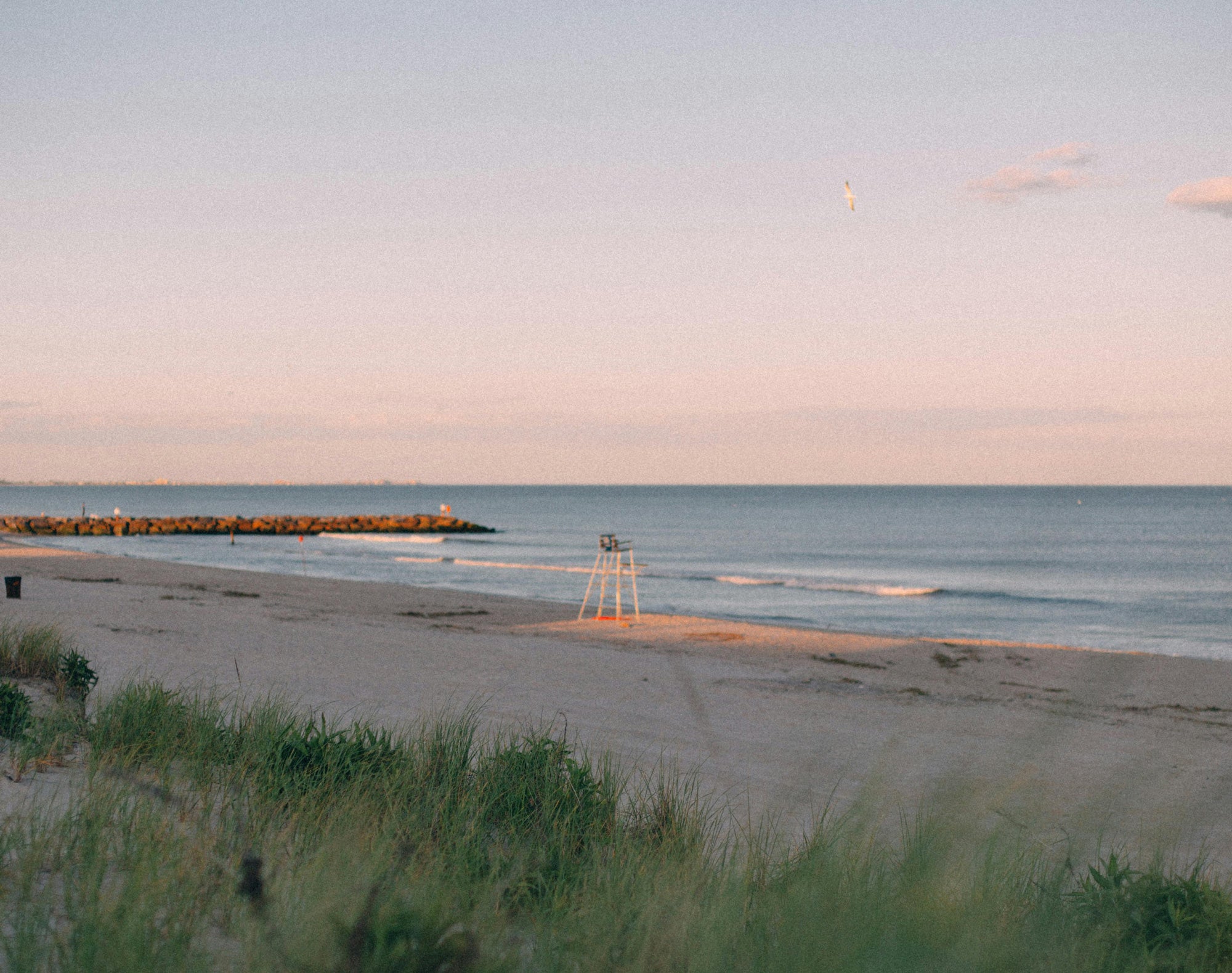 Sunset view of a beach with dune grass, a lifeguard stand, and a stone jetty