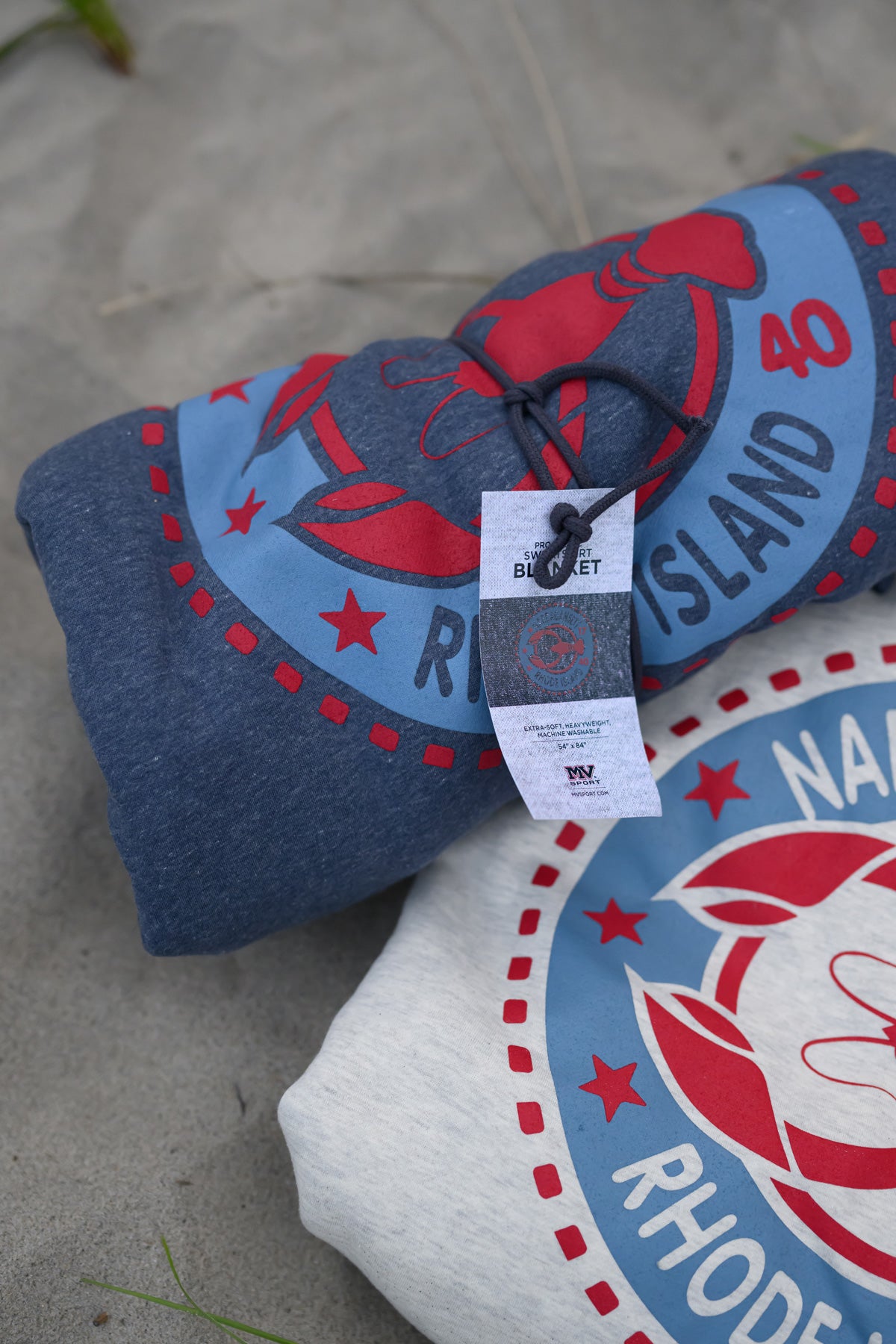 Rolled Rhode Island lobster-print sweatshirt blankets on a sandy Narragansett beach
